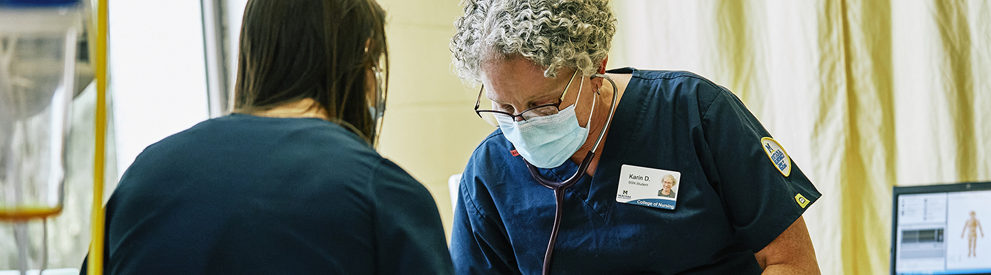 A woman with curly gray hair wearing scrubs and PPE observes a patient.