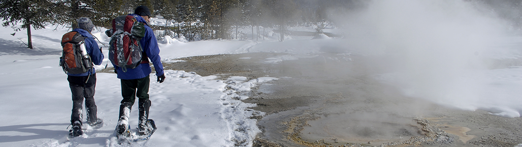 Two people snowshoe past a steaming creek in winter.