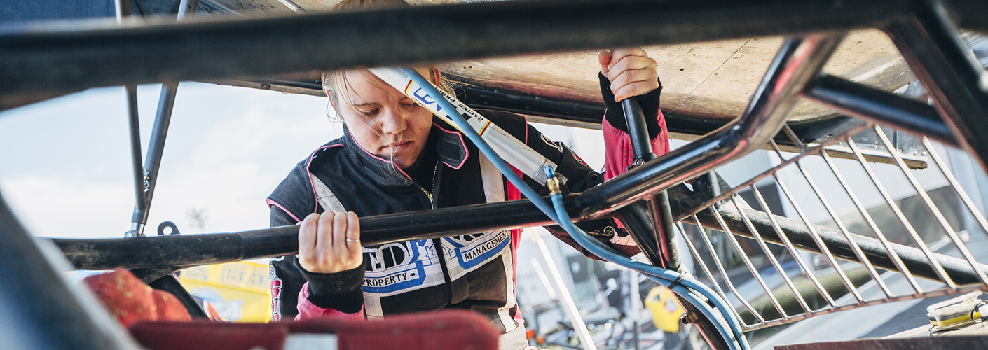 A young woman in racing gear tests the supports on a race car frame.