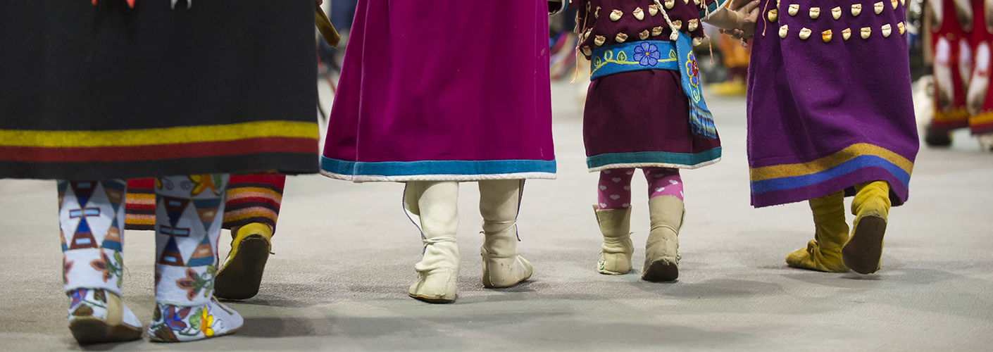 A line of three women and a toddler-aged girl, all in traditional American Indian dress at a pow-wow, walk towards the festivities.