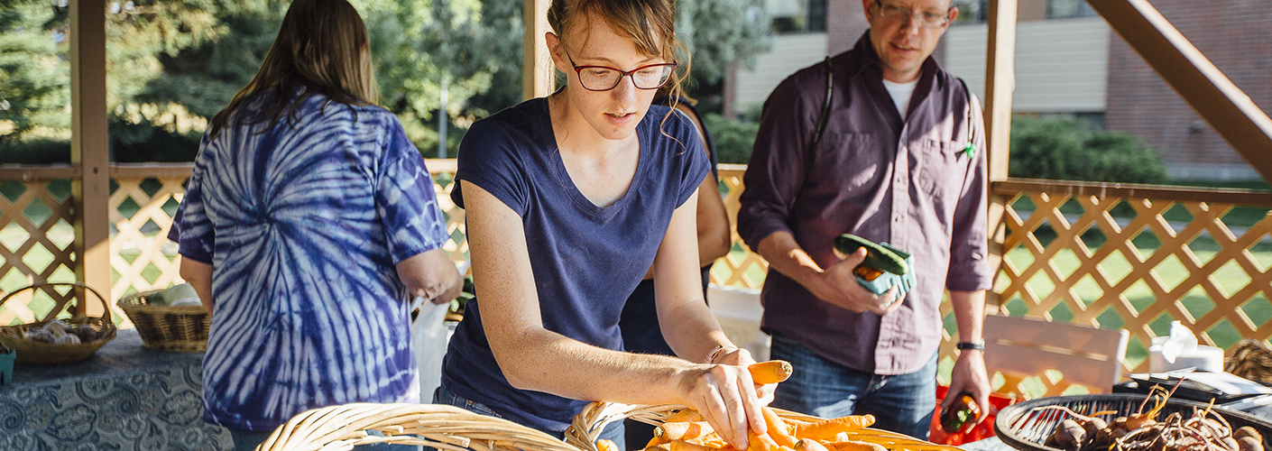 A young woman in glasses arranges carrots in a farmer's market stall, while a man with produce in his hands looks on.