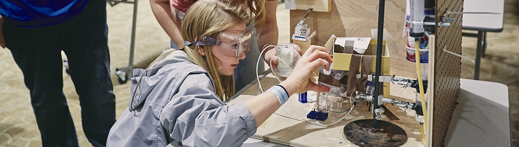A young woman with safety goggles inspects a student's project.
