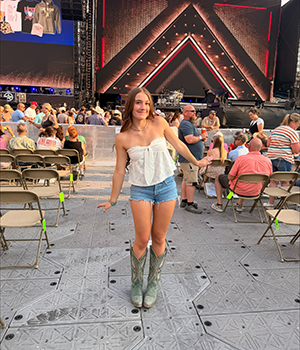 A girl with long brown hair and a white shirt and jean shorts posing at a concert