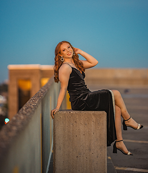 A girl with long curly orange hair wearing a long black dress posing on a pillar