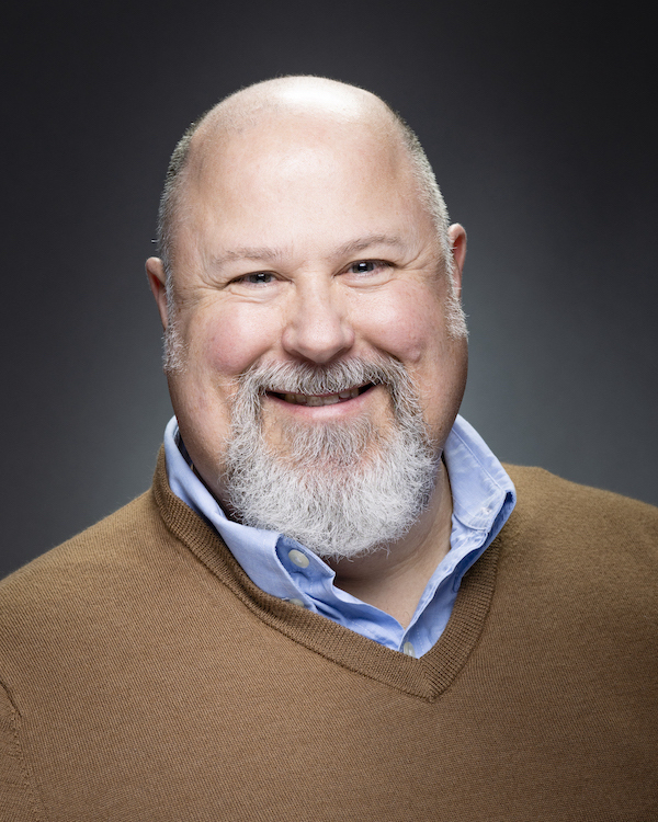 A bald man with a gray beard wearing a brown sweater smiling to the camera