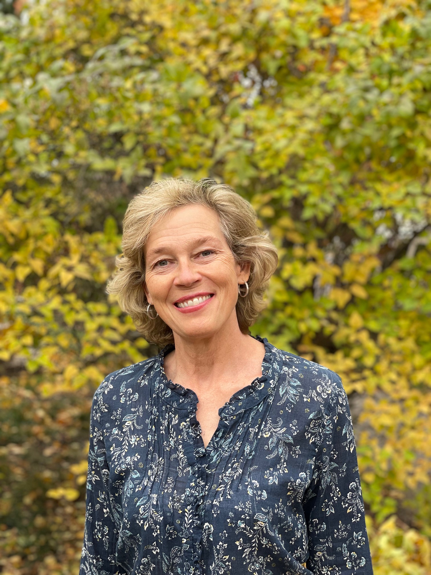 A woman with short hair and a blue floral shirt smiling to the camera with trees behind her