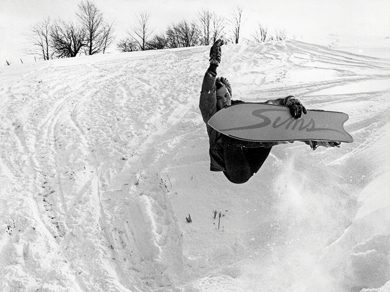 A black and white image of a person mid-air holding their snowboard with one hand