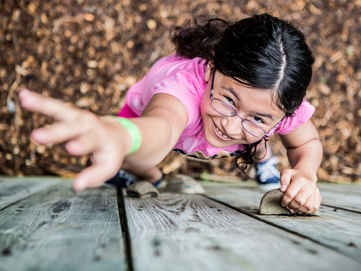 A young girl wearing glasses and a pink shirt climbing up a wooden climbing wall