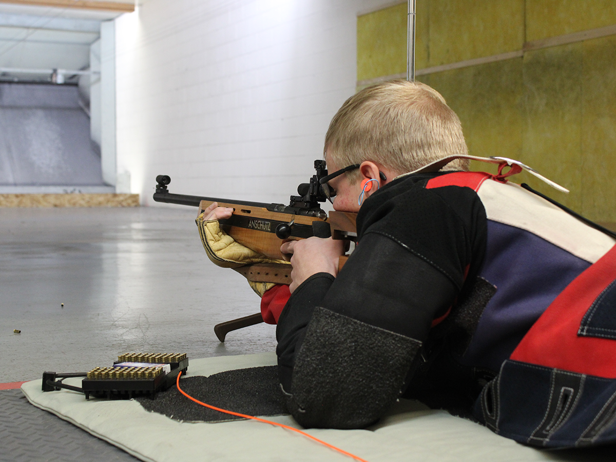 A youth with short blonde hair laying prone pointing a rifle at a target