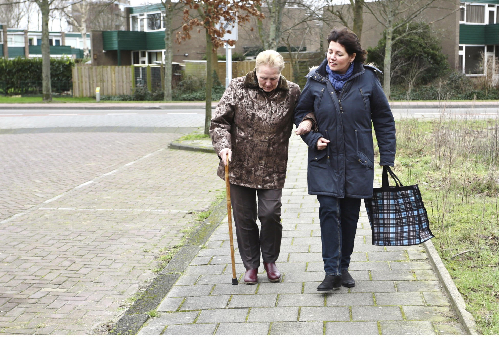 Two people wearing coats with linked arms walking down a stone sidewalk. The person on the left is using a cane and the person on the right is carrying a bag