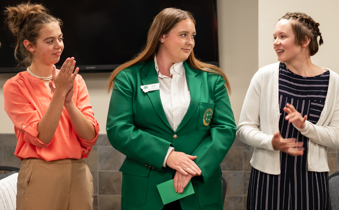 Three girls clap and look at each other; the middle girl has a green 4-H blazer on.