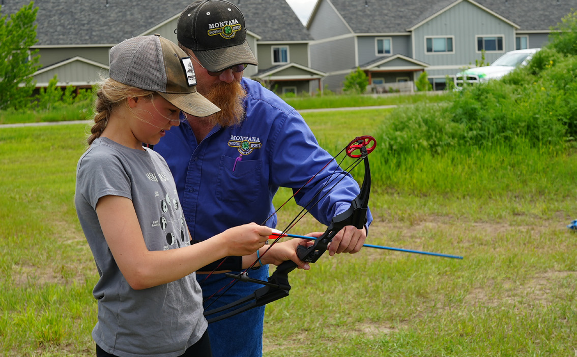 A male figure helps a young girl insert an arrow into a bow.