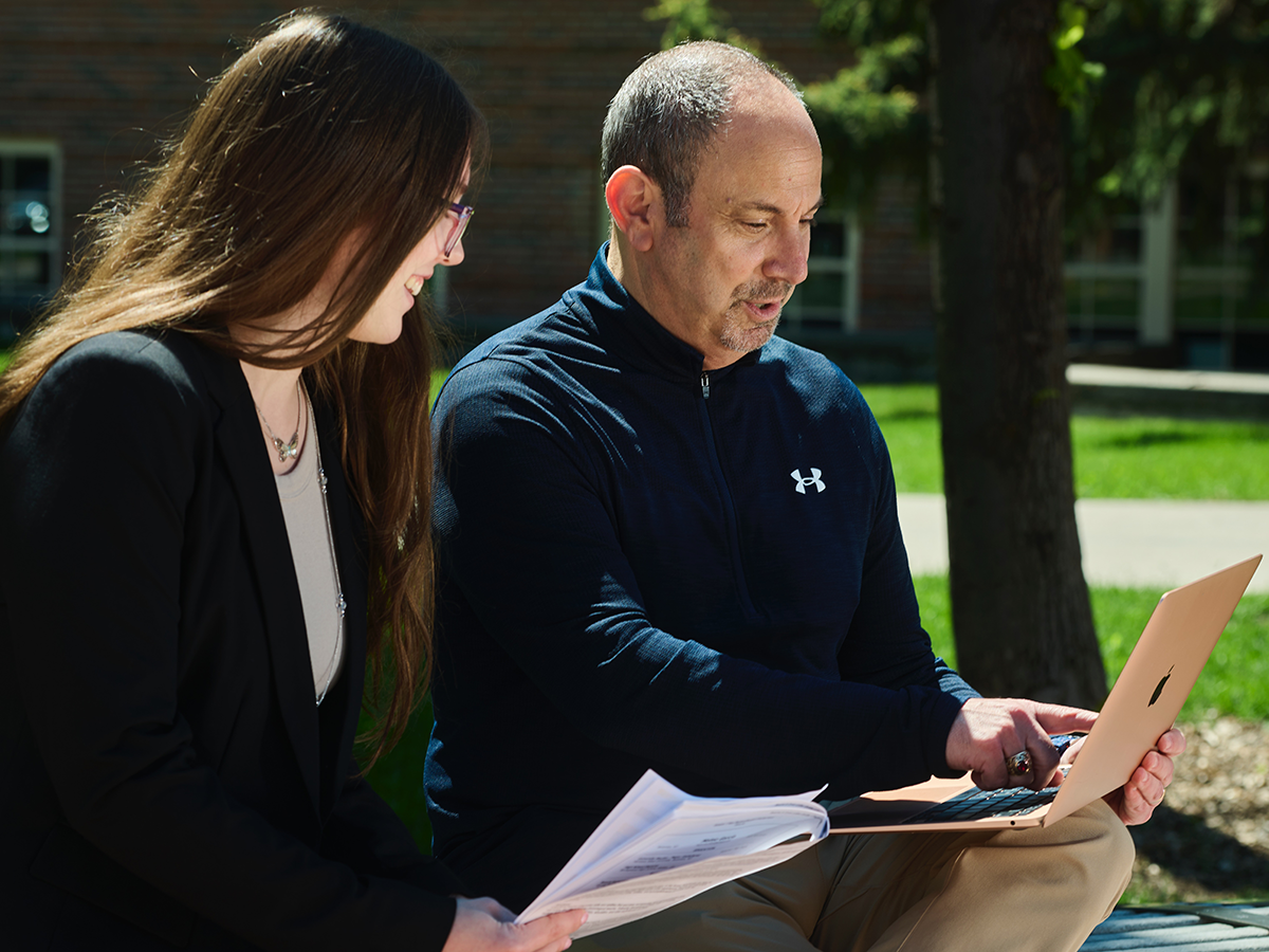 A person with a dark blue zip up sweatshirt pointing at something on a laptop, with another person with long dark hair sitting next to them
