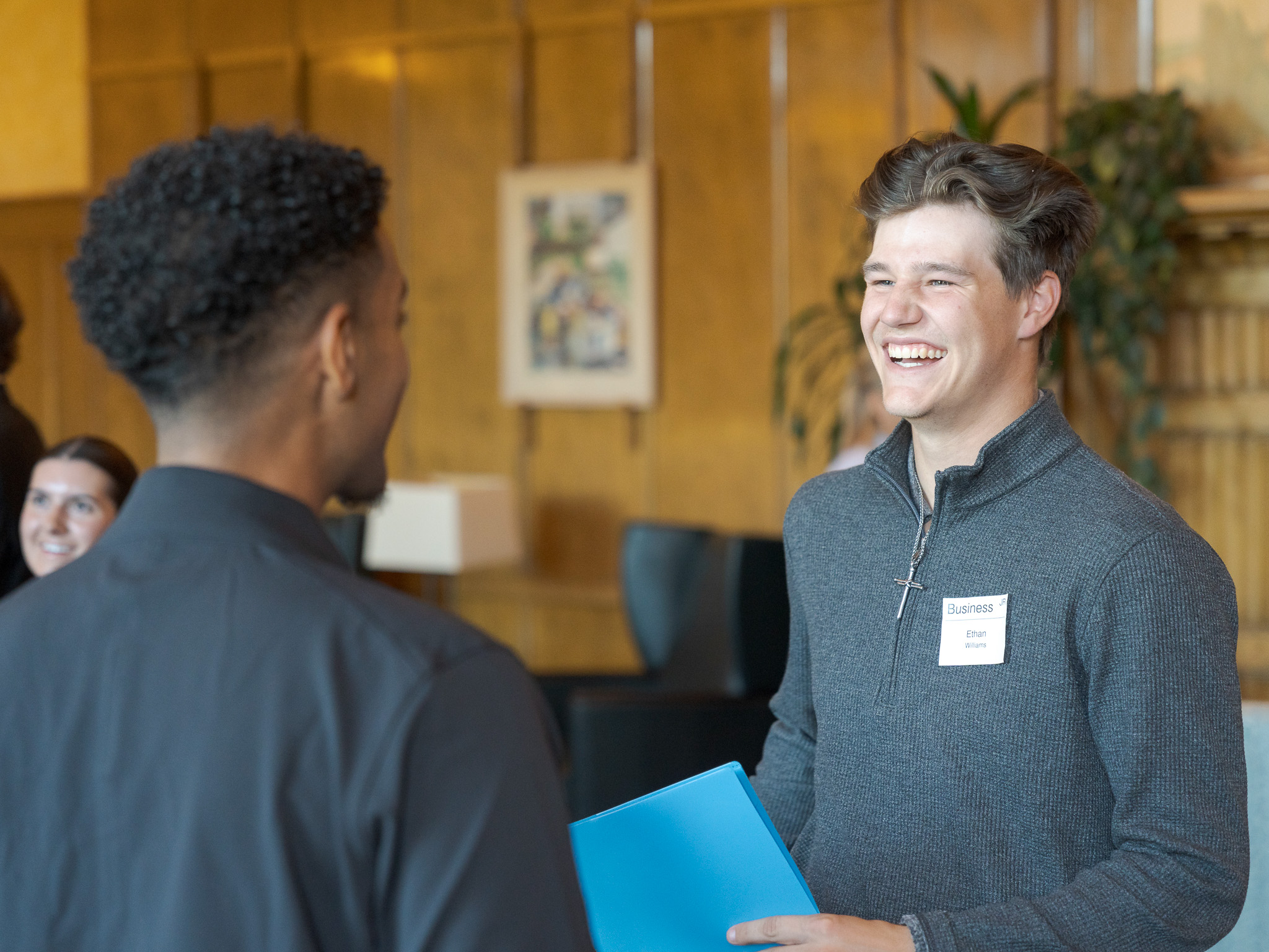 A man wearing a quarter-zip sweatshirt holding a blue paper smiling and talking to another person
