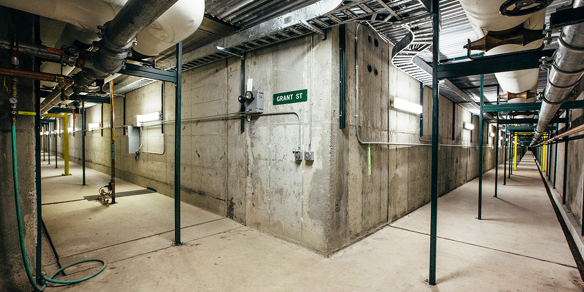 The corner of a hallway with left and right paths. Concrete walls floors with pipes along them.